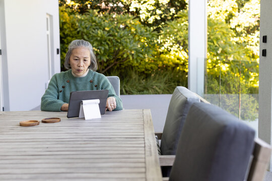 Senior Asian woman sitting at patio table wearing green sweater using tablet on stand, copy space