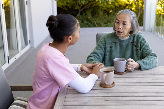 Asian senior woman and caregiver in pink scrubs holding hand, chatting over mugs on balcony table