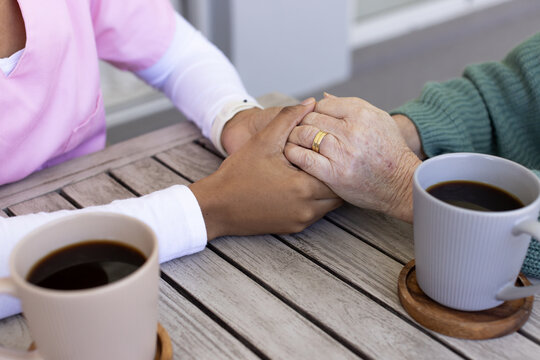 Female senior Asian and African American aide holding hands on wooden table with mugs showing ring