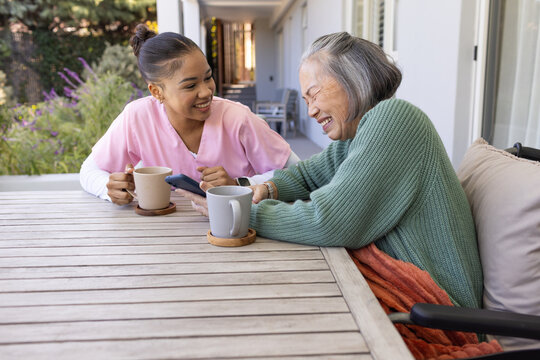 Diverse female caregiver showing smartphone and mug while senior female laughing on covered patio