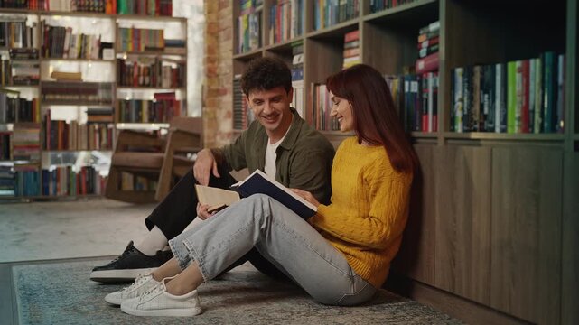 Red-haired woman and man sitting in library aisle, reading books together