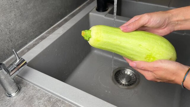 Close up of female hands washing fresh green zucchini under running water in modern kitchen sink. Food preparation, hygiene, and healthy cooking concept