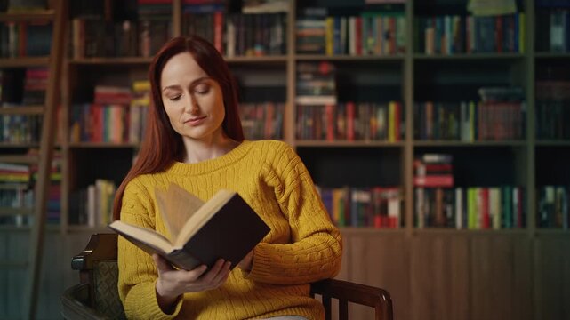 Relaxed young red-haired woman reading a book in urban library, enjoying hobby