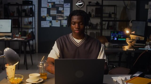 Front view medium shot of African American young adult man working on laptop computer at office desk at night, reaching for drink. For commercial projects about overtime work, corporate productivity