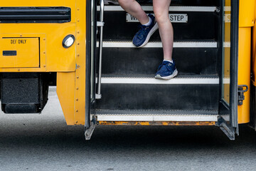 Child in navy sneakers steps down from a yellow school bus onto the pavement, capturing the everyday transition between home and school while illustrating independence and back-to-school. © Jeanne Sager Photo