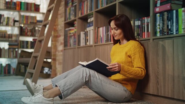 Relaxed young female student reading a book on floor in academic library