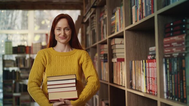 Portrait of young red-haired woman holding stacks of books in bookshop
