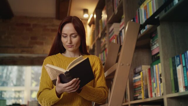 Positive young woman reading a book on library ladder, enjoying favorite hobby