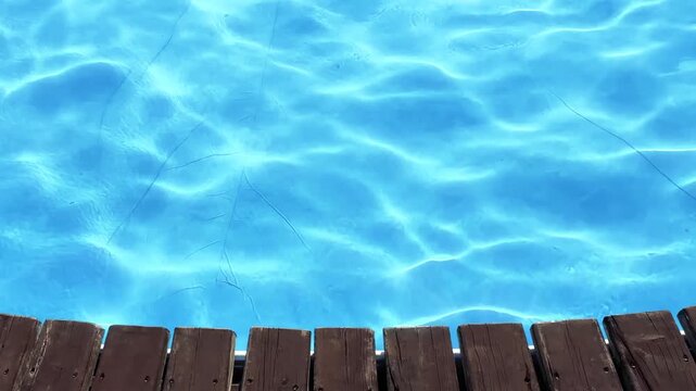 Sunlight reflecting on clear blue swimming pool water with gentle ripples next to a wooden deck. Perfect background for summer vacation, resort, and relaxation concepts