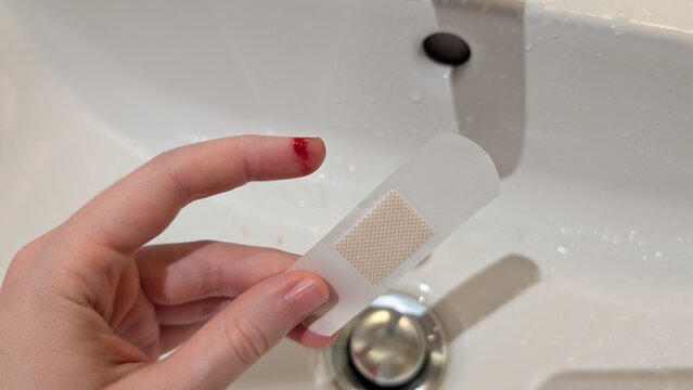 Domestic accident concept close-up of a hand with a bleeding cut index finger and an adhesive bandage held between fingers over a white sink.