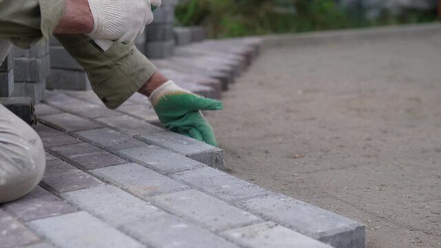 Close side view shows worker hands in protective gloves setting concrete pavers along straight walkway edge with rubber mallet, partial body at left, blurred site background, and