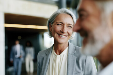 Obraz premium A joyful woman with gray hair beams with happiness in a corporate setting while engaging in a candid conversation with colleagues, showcasing her vibrant professional persona.