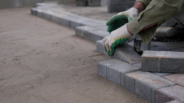 Worker hands in protective gloves set gray paving blocks along unfinished outdoor walkway at right side, forming straight curb edge beside dusty ground, rough sand, stacked stones
