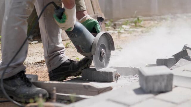 Close-up outdoor work scene shows worker using handheld angle grinder to cut concrete pavers beside dusty ground, with gloved hands, sturdy black shoes, scattered stone blocks