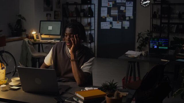 Wide shot of African American young adult man working late on laptop in office, holding head in hands from stress and fatigue. For corporate mental health and workplace burnout marketing campaigns
