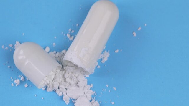 Top view Close-up macro shot of a white medicine capsule opening and releasing powder, rotating slowly against a blue background.