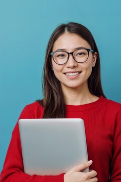 Young Asian woman smiles warmly while holding a laptop on blue background. She wears glasses and a cozy red sweater