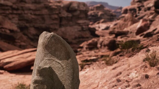 A stone monolith rises from the ground in a rocky canyon. Sunlight shines on the rock formations. The landscape is dry with some sparse vegetation.