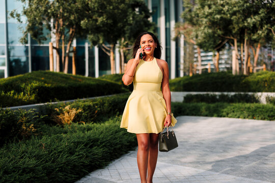 Smiling woman in yellow dress walking outdoors, communicating on her cell phone in a modern urban park setting