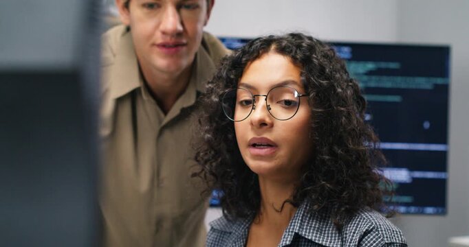 Hispanic man and woman programmers pair on code review across monitors focusing on security database integration ai features and product development in a corporate workspace.