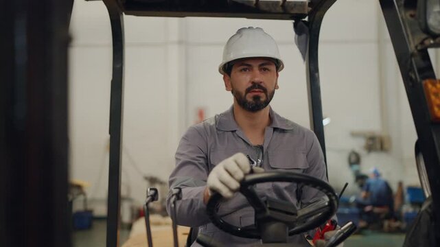 Man wears hard hat, grips forklift controls. Forklift operator rests hand on steering wheel. Factory background holds tools and storage racks. Determined look shows focus and readiness