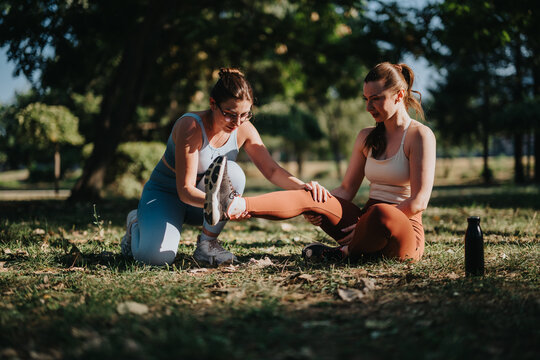 A personal trainer helps a woman stretch her leg while seated on the grass. Two women perform a guided hamstring stretch together during an outdoor workout session.