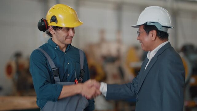 Two men shake hands in a factory setting. One wears a suit, the other overalls and a hard hat. They stand near large machinery in an industrial space