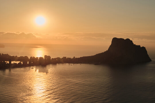 Golden hour over a beautiful coastal city, with the sun casting a brilliant glow across the tranquil ocean. The iconic Penon de Ifach rock stands majestically against the warm, serene sky.