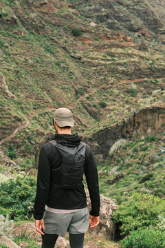 Rear view of a man in outdoor gear with a minimalist backpack, looking out over a vast, green, and rugged mountain landscape with visible winding trails, evoking adventure and exploration.