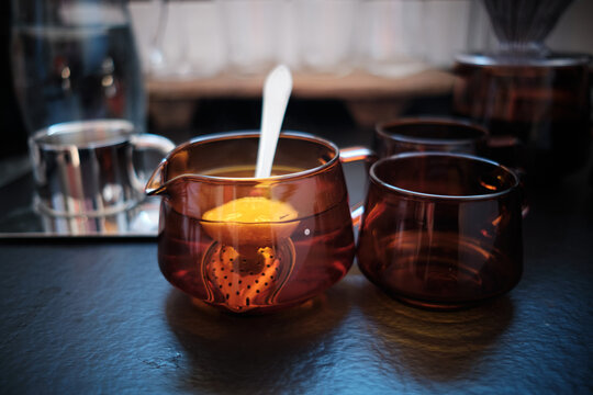 Close-up of a stylish amber glass teapot brewing aromatic tea with a fresh lemon slice and infuser, accompanied by matching cups on a dark table. Perfect for relaxation and cozy moments.