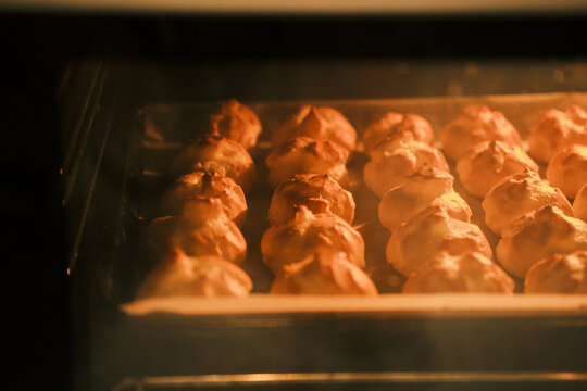 Freshly baked golden choux pastry puffs rising in a warm oven, creating a comforting and appetizing scene of homemade dessert preparation. Perfect for food blogs, recipes, or culinary themes.