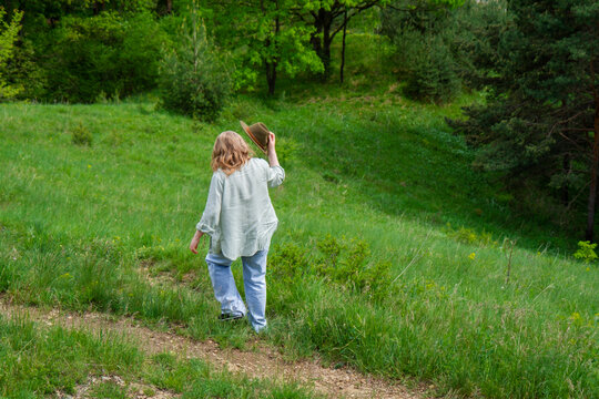 Back view of woman holding hat walking down dirt path on green grassy hill slope. Concept of solo travel, tranquility and escaping city.