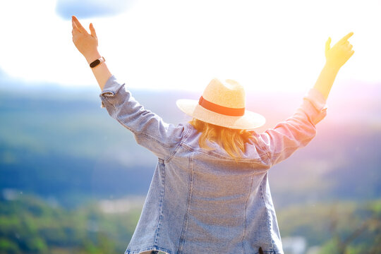 Blonde woman in denim jacket raising hands towards sun in mountains from behind. Harmony with nature, spiritual freedom, outdoor adventure, and positive mind concept.