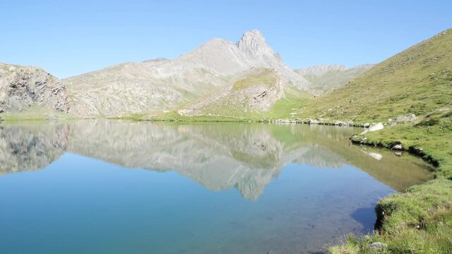 Crystal clear alpine lake reflecting rugged mountain peaks beneath cloudless blue sky in Italian Alps, showcasing pristine wilderness, tranquil waters and spectacular summer scenery