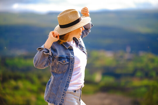 Joyful blonde woman in straw hat adjusting hat brim in nature. Summer travel, outdoor recreation, active lifestyle, and positive energy concept.