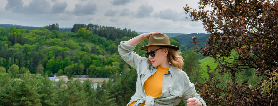 Stylish blonde woman in green hat and sunglasses standing against green mountain forest landscape. Summer travel, eco tourism, and outdoor leisure concept.