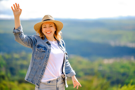 Laughing blonde woman in straw hat waving hand outdoors in sunny weather. Positive mood, summer vacation, outdoor recreation, and friendly greeting concept.