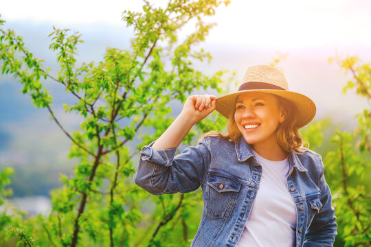 Smiling blonde woman in denim jacket touching straw hat under green tree outdoors. Summer beauty, harmony with nature, positive energy, and active lifestyle concept.