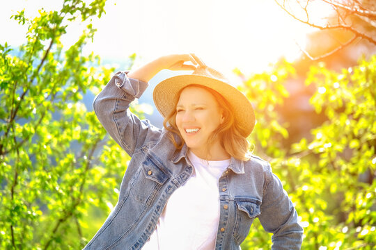 Radiant blonde woman in straw hat touching brim under bright sunlight outdoors. Summer beauty, positive mood, and harmony with nature concept.