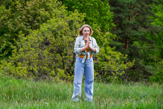 Calm blonde woman meditating with hands in prayer pose in green forest meadow. Yoga outdoors, mental peace, mindfulness, and spiritual wellness concept.