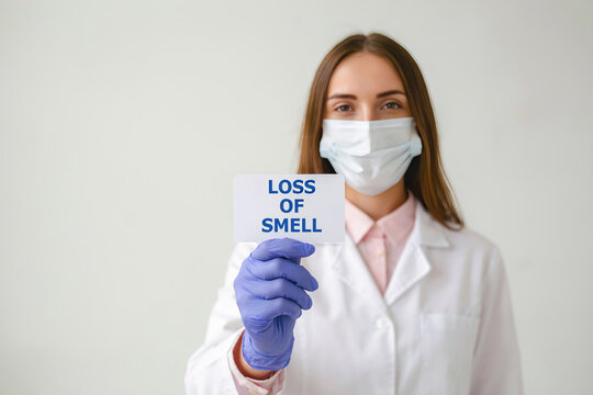 Woman doctor wearing mask and gloves holding card with 'Loss of Smell' during medical consultation