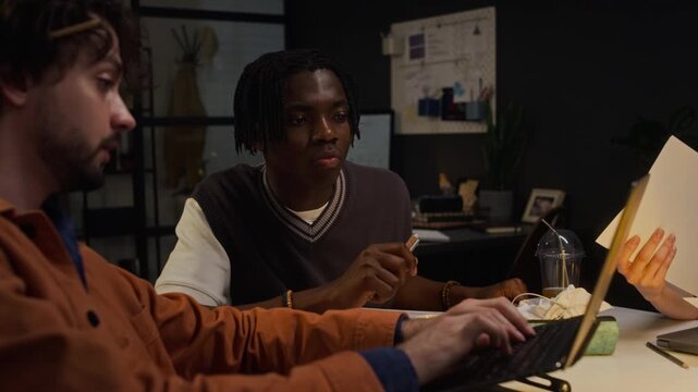 Caucasian young adult man typing on laptop while African American young adult colleague pointing at screen, discussing project late in office. Video suitable for corporate teamwork presentations
