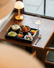 High-angle shot of a complete Japanese bento lunch set served on a tray with a glass of white wine and a modern gold desk lamp on a wooden table near a window. © Simone Oppes
