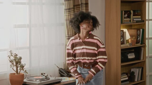 African American young woman playing vinyl record and dancing energetically while Caucasian young friends watch from couch. Ideal for retro lifestyle and music streaming service commercials