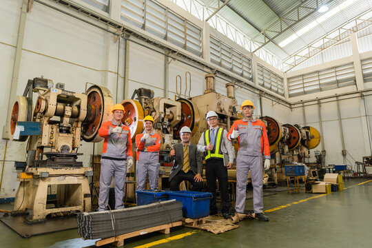 Confident factory team gives thumbs up beneath tall industrial roof near press machines