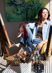 Portrait of a female artist relaxing beside her easel in a bright studio filled with warm natural sunlight. © Diana