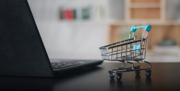 Small shopping cart on office desk next to laptop computer. Concept of online shopping, e-commerce business, digital retail marketing and consumer purchase behavior with copy space background.