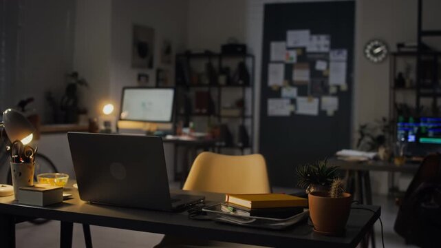 Panning shot of empty modern office workspace showing open laptop on desk at night. Clip ideal for corporate presentations, business marketing, or overtime work concepts