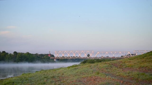 View of a railway bridge crossing over a calm river with a passenger train passing through on an early foggy morning