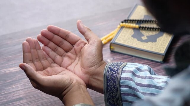 A close up shot of a man's hands as he prays, holding them open in devotion with a holy book and prayer beads in the background, offering a serene and spiritual moment captured in slow motion